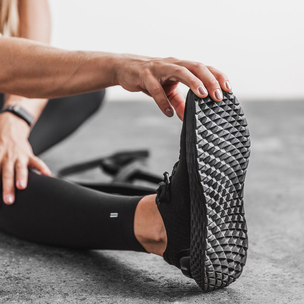 A woman wearing athletic apparel stretches before a workout wearing black ripstop runner training sneakers