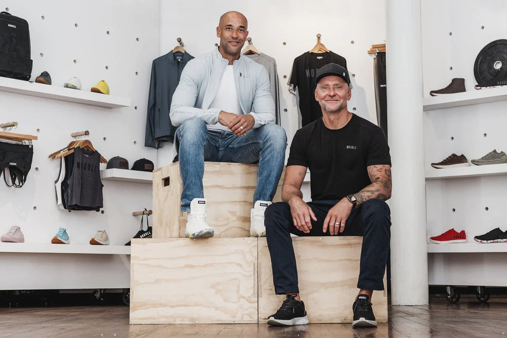Two men wearing casual clothes sit on boxes while surrounded by NOBULL apparel and workout gear