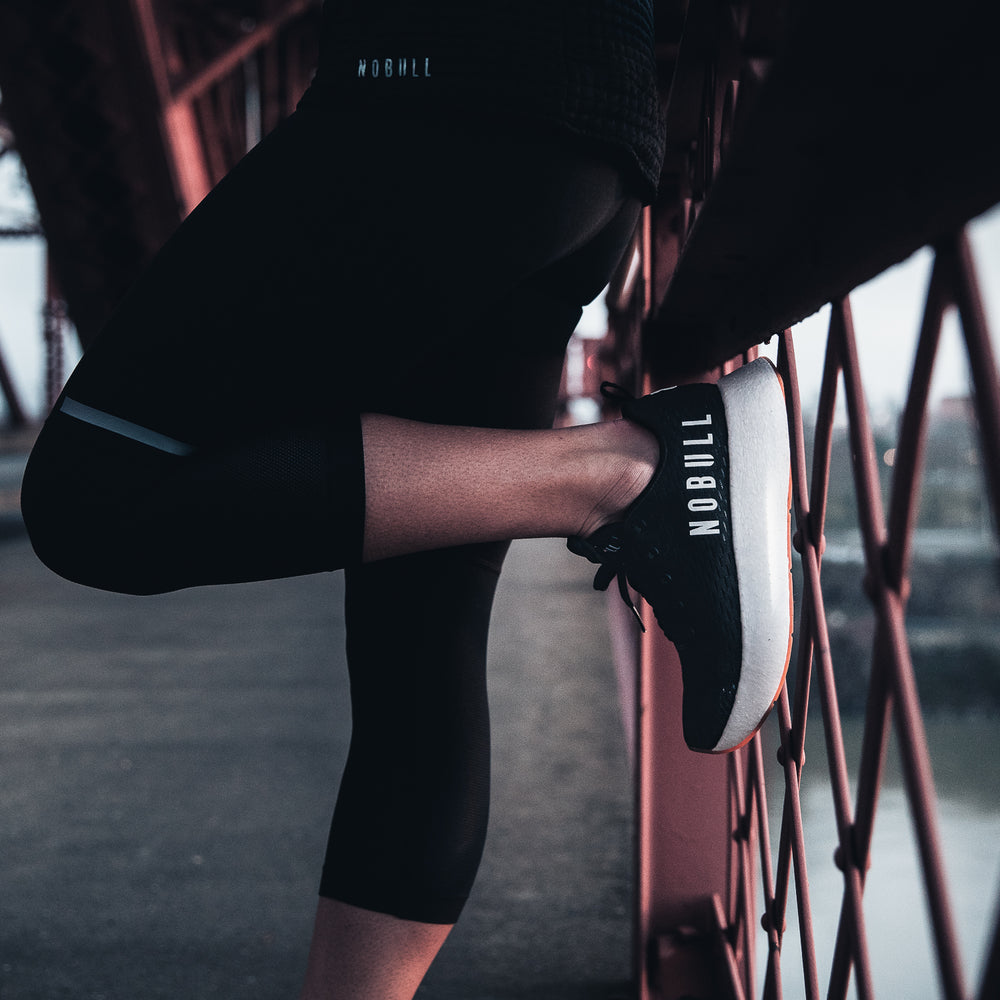 A women wearing athletic clothing and tights stands on a bridge wearing black and white runner plus shoes