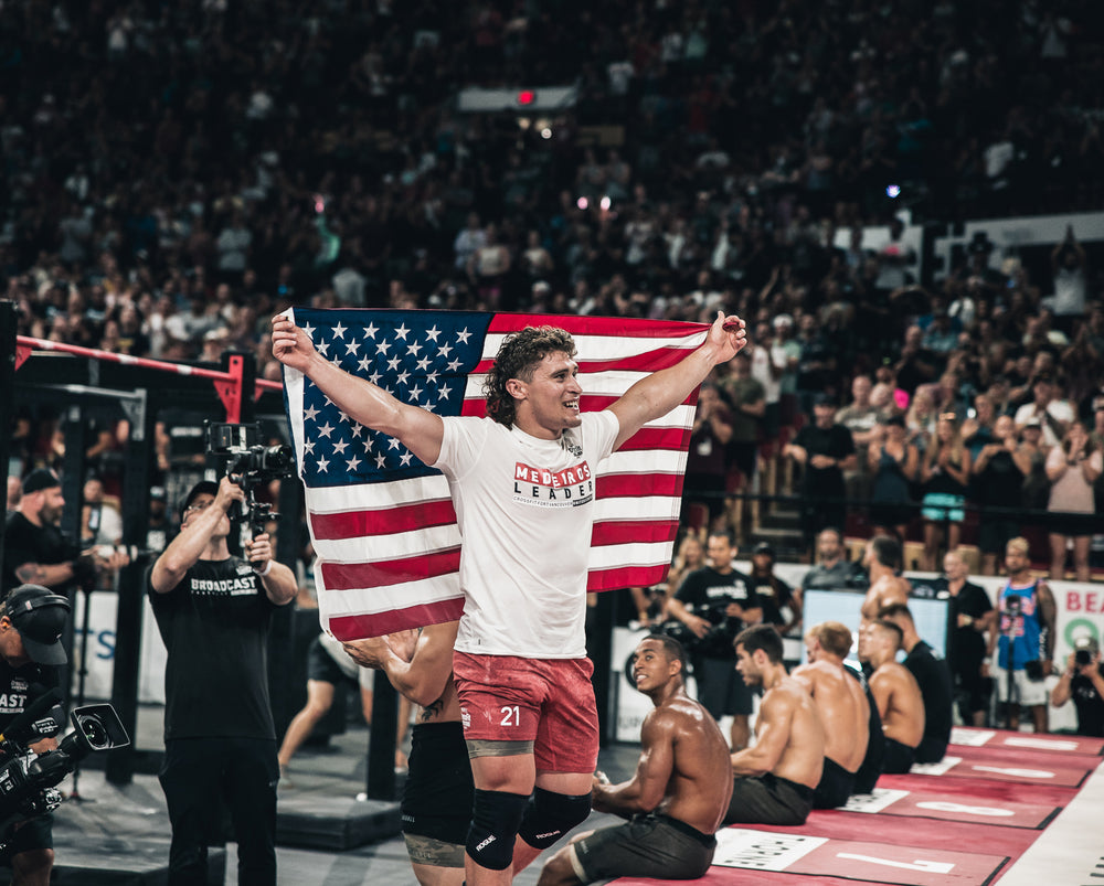 CrossFit Games Champion Justin Medeiros wears athletic clothing and holds an american flag behind him