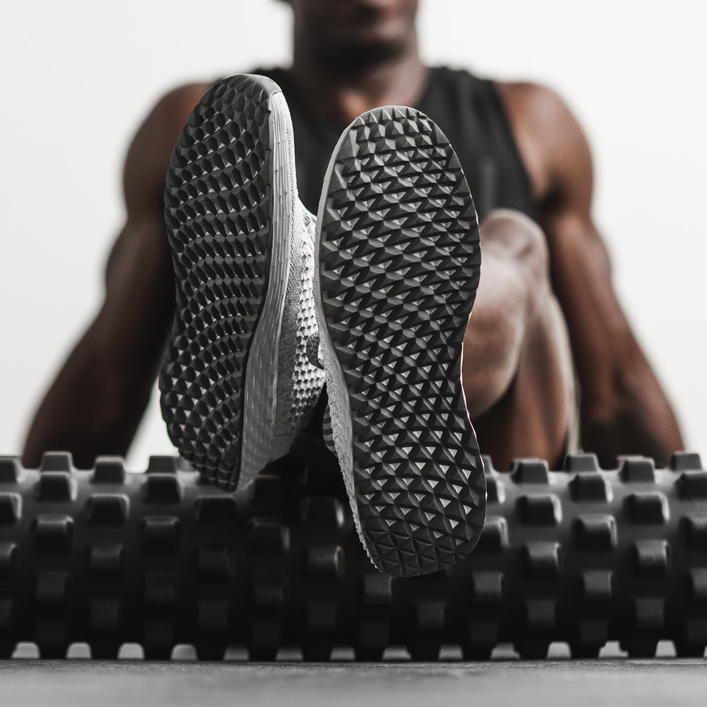 A man in workout apparel and grey knit training shoes uses a foam muscle roller